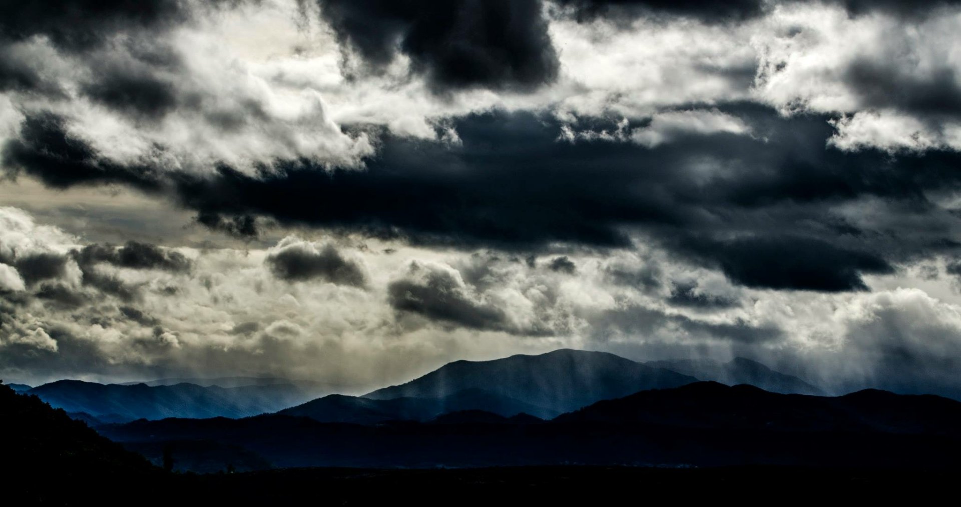 HD desktop wallpaper showcasing a dramatic mountain landscape under dark clouds with sunbeams breaking through, highlighting the natural beauty and moodiness of the scene.