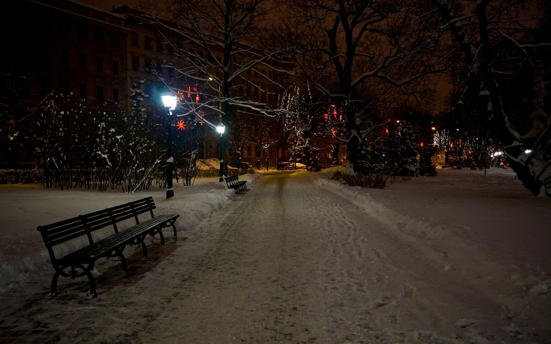HD desktop wallpaper of a snowy park at night featuring man-made benches along a snow-covered pathway illuminated by streetlights and festive lights.