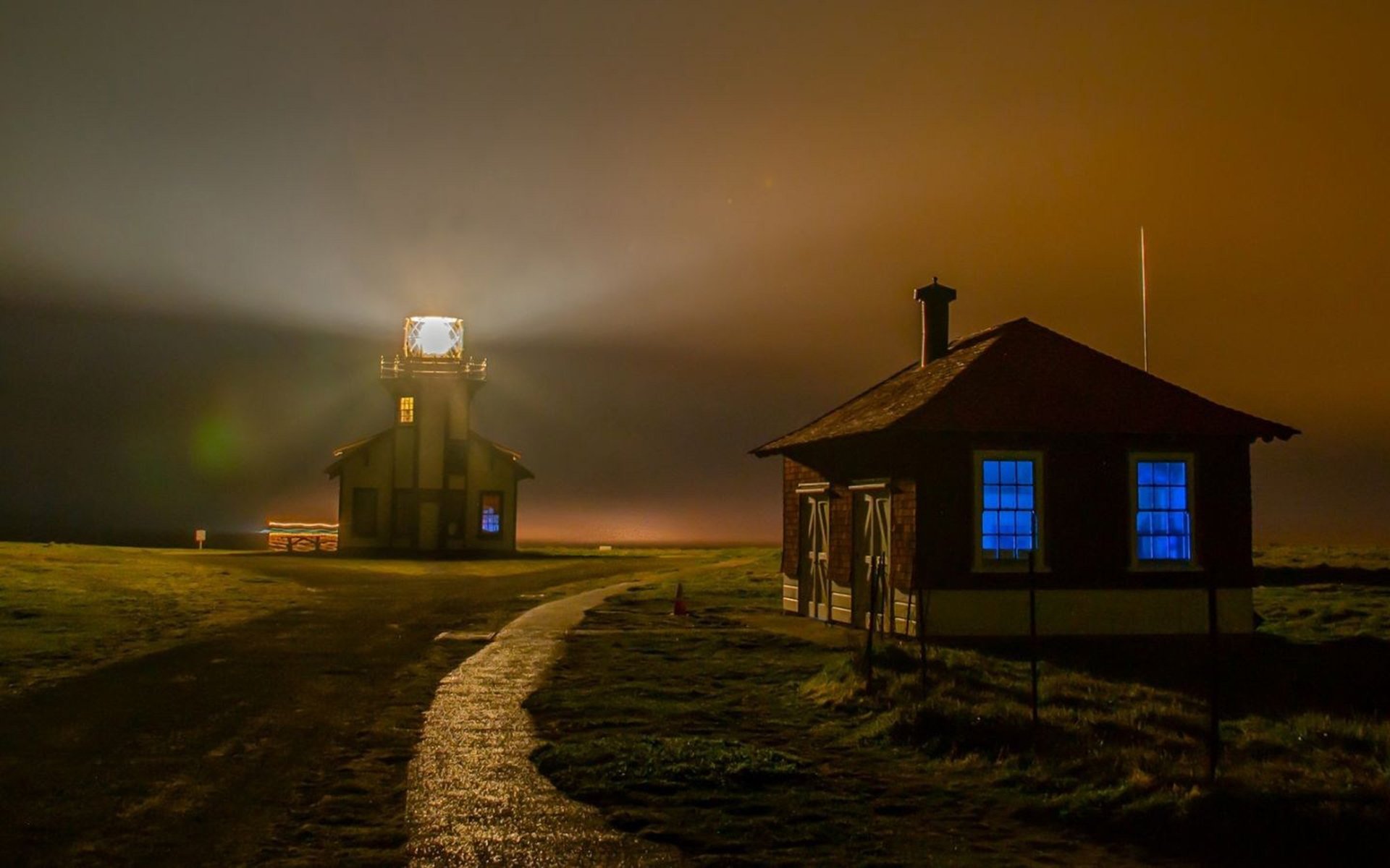 HD PC desktop wallpaper and background: man-made coastal night scene with an illuminated lighthouse casting beams across mist, and a small house with blue-lit windows by a winding path.