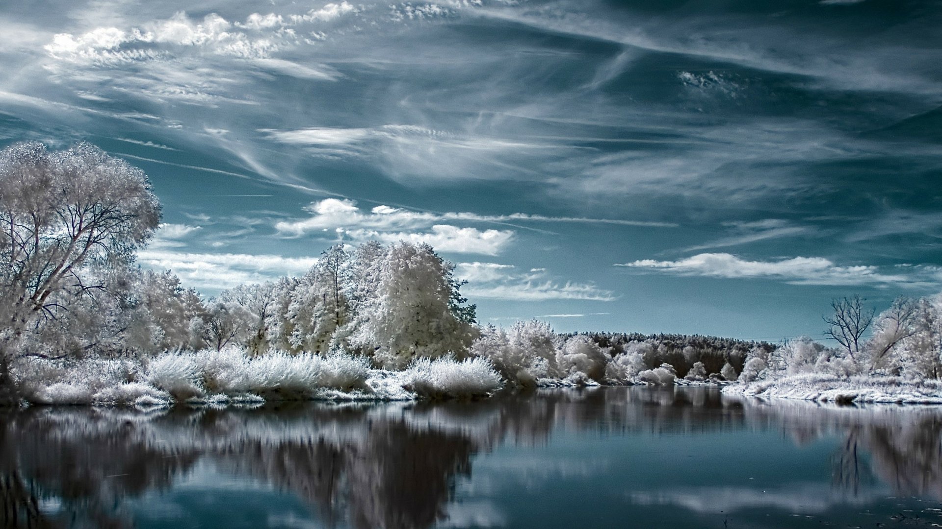 A serene winter scene with a tranquil lake surrounded by snow-covered trees under a partly cloudy sky. This HD wallpaper captures the beauty of nature in winter.