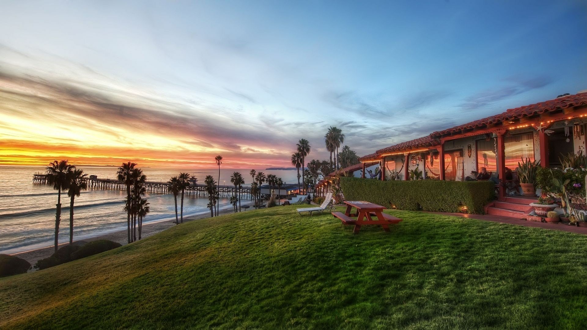 HD PC desktop wallpaper and background: man-made cottage on a grassy coastal bluff with palm trees overlooking the ocean at sunset.