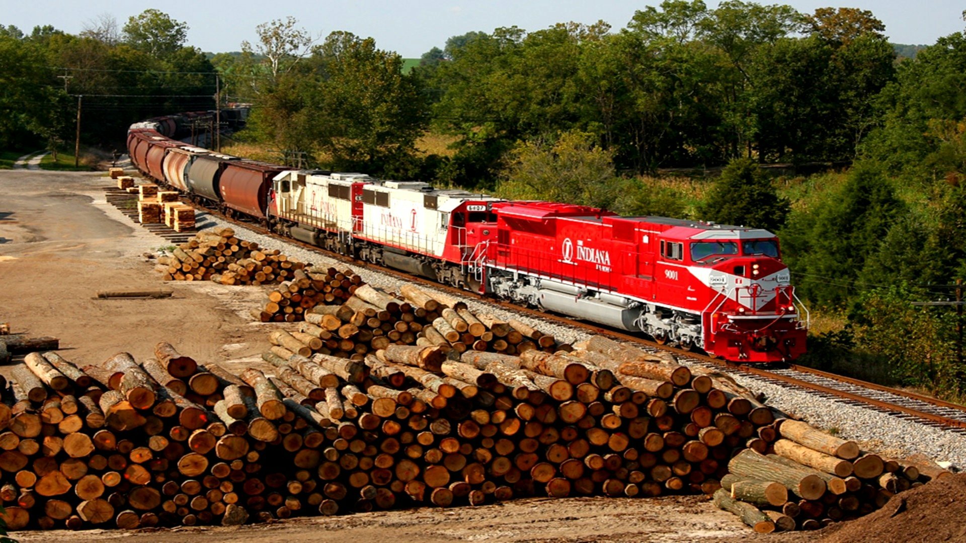 A vibrant red freight train travels along tracks beside stacked logs in a forested area, captured in an HD desktop wallpaper showcasing a vehicle in motion.