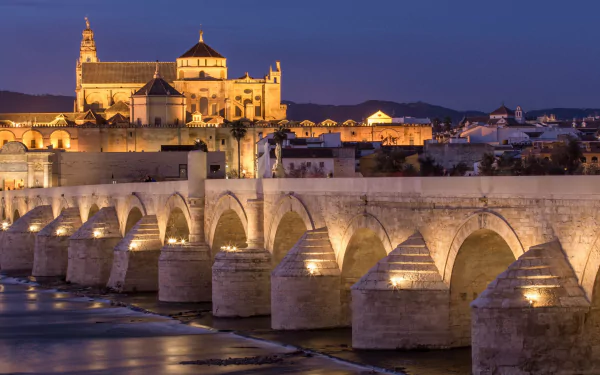 A stunning view of the Roman Bridge of Córdoba, illuminated at dusk, showcasing its arches and the historic architecture of the surrounding city. 4K Ultra HD background image.