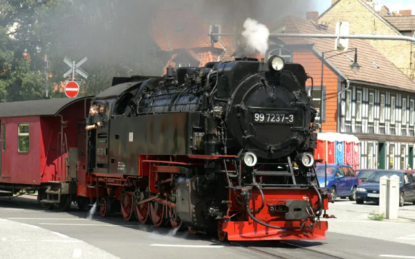 HD PC desktop wallpaper and background: black-and-red steam train vehicle steaming through a small town, puffing white smoke as it pulls a red passenger car.