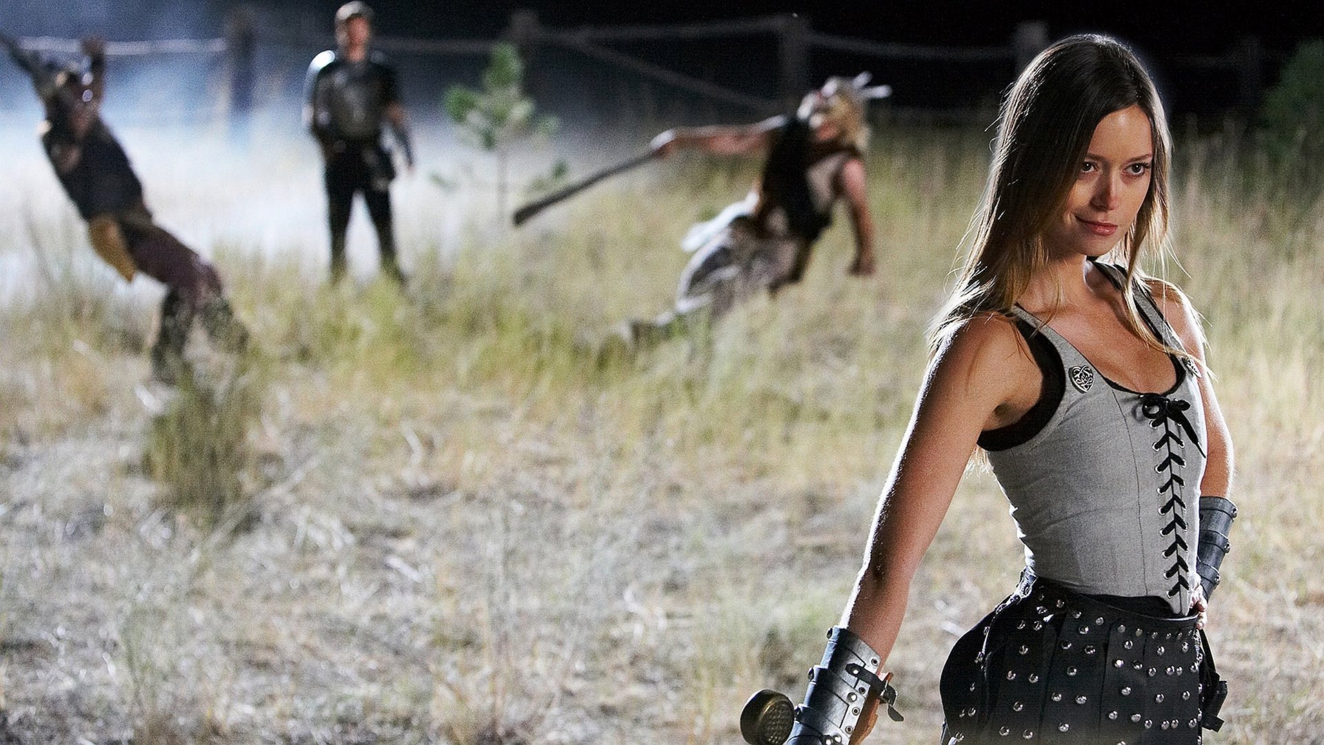 Movie 'Knights of Badassdom' HD PC desktop wallpaper/background: corseted warrior woman with arm bracer and sword stands in a moonlit field as blurred costumed fighters clash behind her.