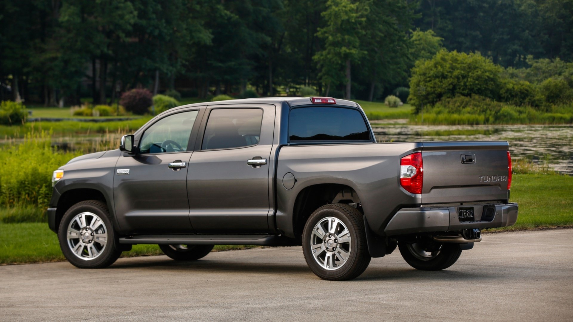 HD PC desktop wallpaper of a grey Toyota Tundra pickup truck parked by a lakeside with lush green trees and lawn in the background.