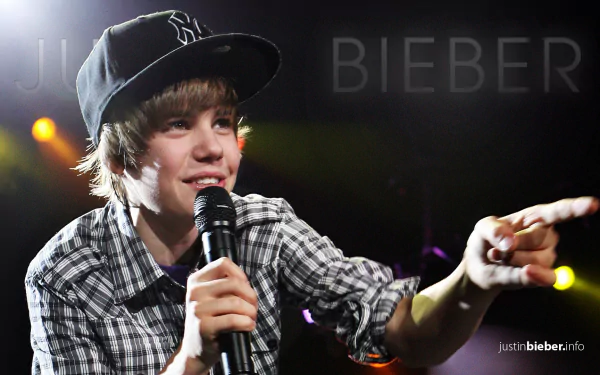 Young male artist performing live with a microphone, wearing a baseball cap and plaid shirt, with stage lights in the background, as HD desktop wallpaper.