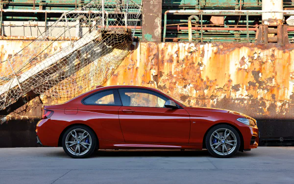 Side view of a red BMW 2 Series coupe parked against a rusted industrial wall — 2K Quad HD PC desktop wallpaper background showcasing the vehicle.