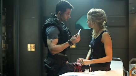 Scene from the movie Lockout shows a man in tactical gear holding an item while talking to a woman tied to a table, presented as a 4K Ultra HD PC desktop wallpaper.