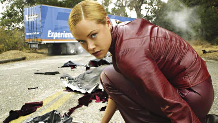 A scene from Terminator 3: Rise of the Machines, featuring a woman in a red outfit crouching on a road, surrounded by debris, with a truck in the background. HD desktop wallpaper.