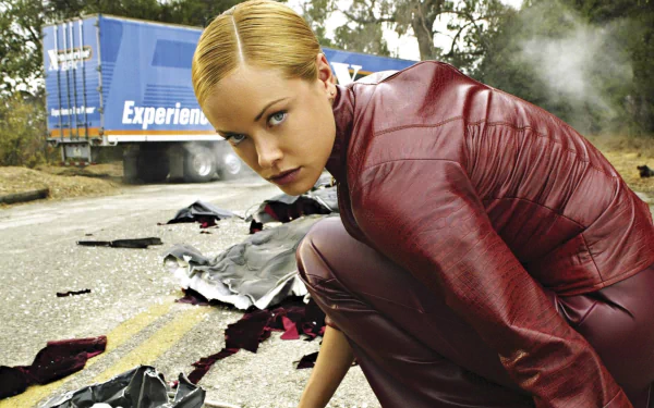 A scene from Terminator 3: Rise of the Machines, featuring a woman in a red outfit crouching on a road, surrounded by debris, with a truck in the background. HD desktop wallpaper.