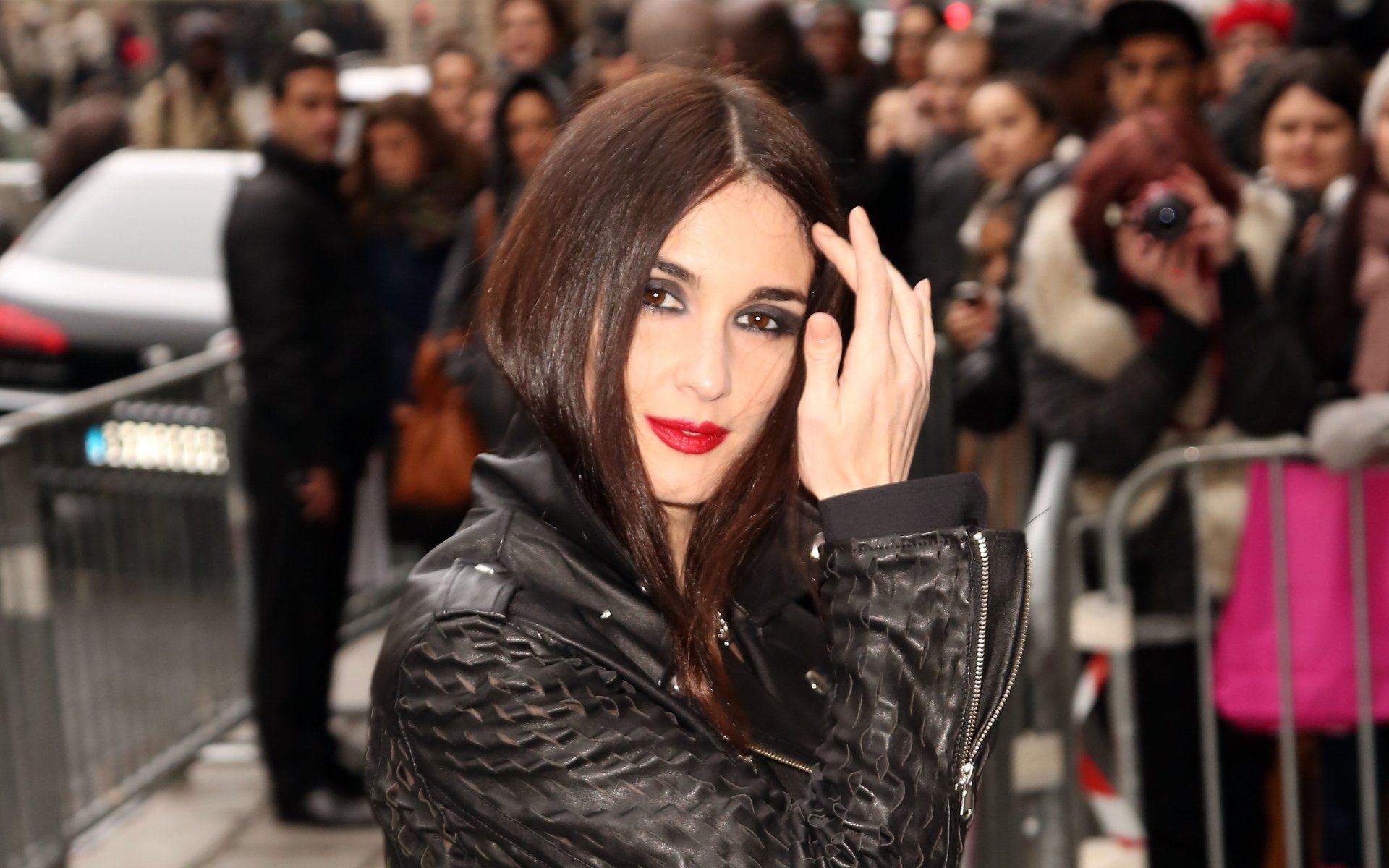 HD desktop wallpaper featuring Spanish actress and celebrity Paz Vega posing confidently in a black leather jacket with a crowd in the background.