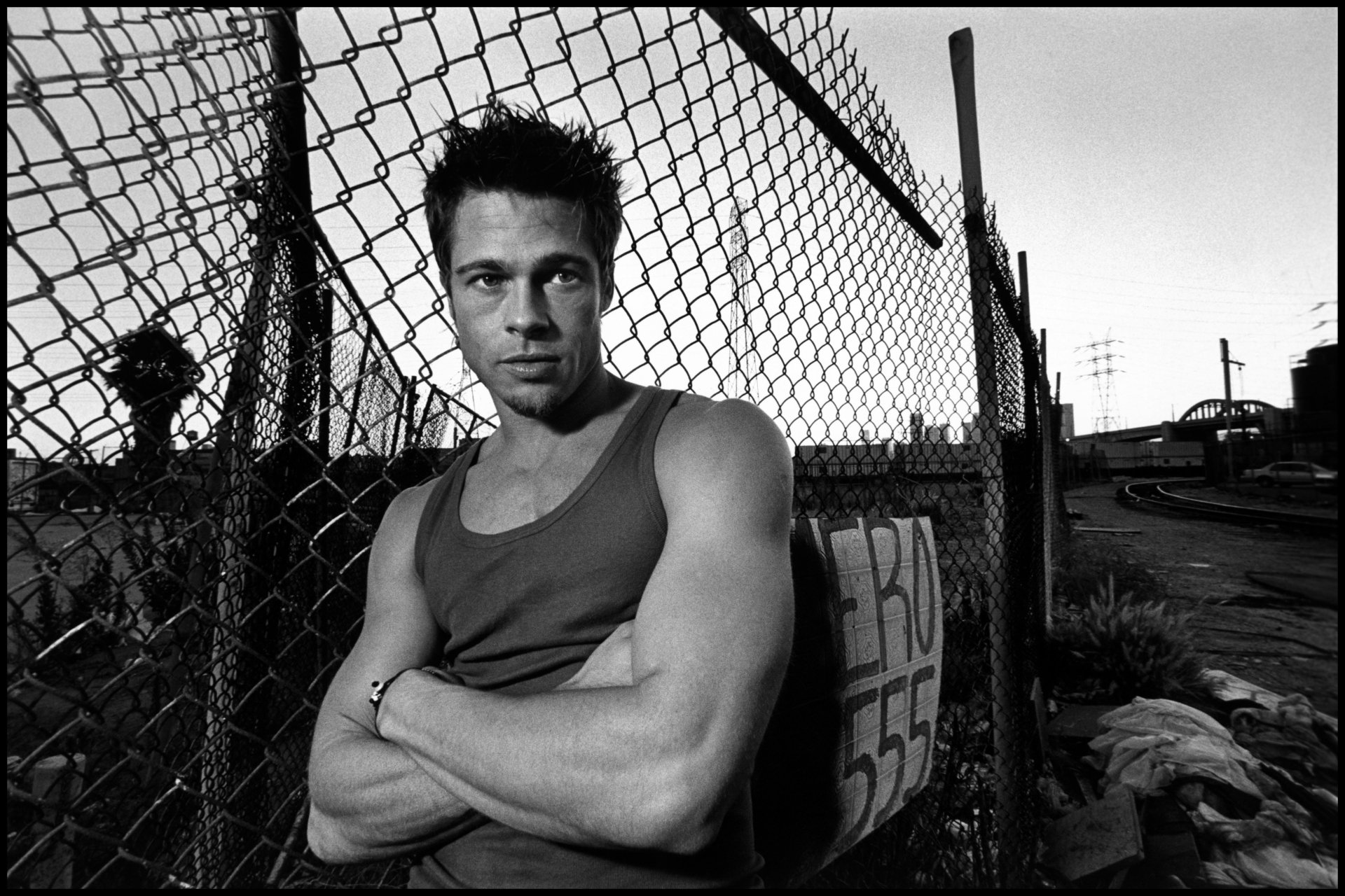 Black-and-white 5K desktop wallpaper of a muscular man in a tank top with arms crossed, leaning on a chain-link fence in a gritty urban landscape.