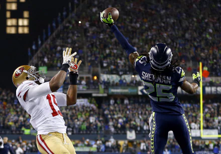 Seattle Seahawks player defends against the opposing team's player, reaching high to intercept a pass in a packed stadium. This HD desktop wallpaper captures the intensity of the game.