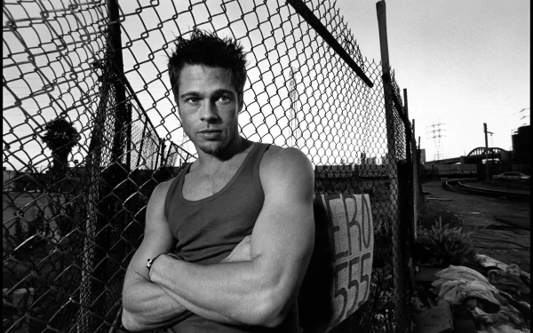 Black-and-white 5K desktop wallpaper of a muscular man in a tank top with arms crossed, leaning on a chain-link fence in a gritty urban landscape.