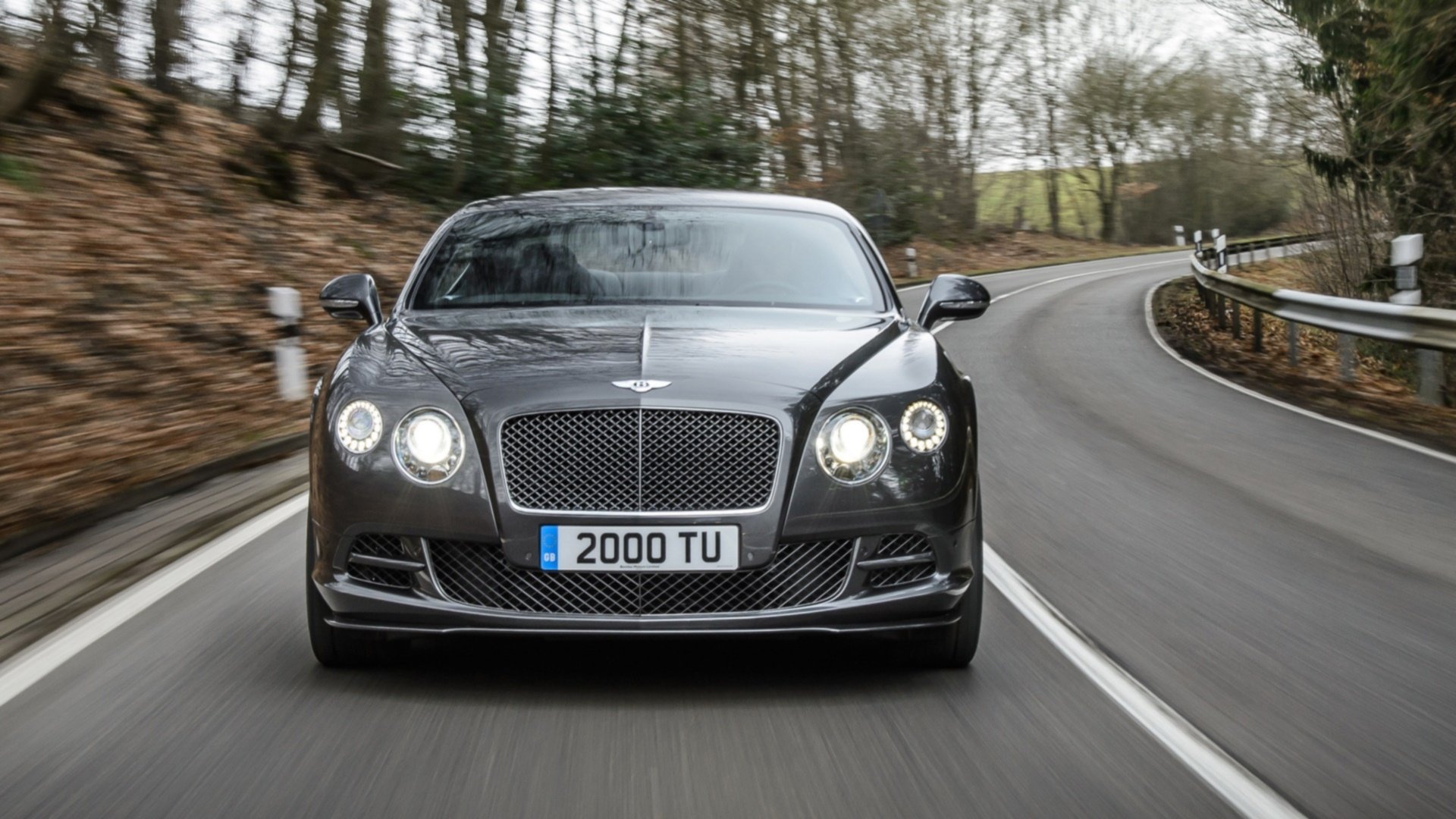 HD PC desktop wallpaper featuring a sleek black Bentley Continental GT Speed driving on a winding road surrounded by trees.