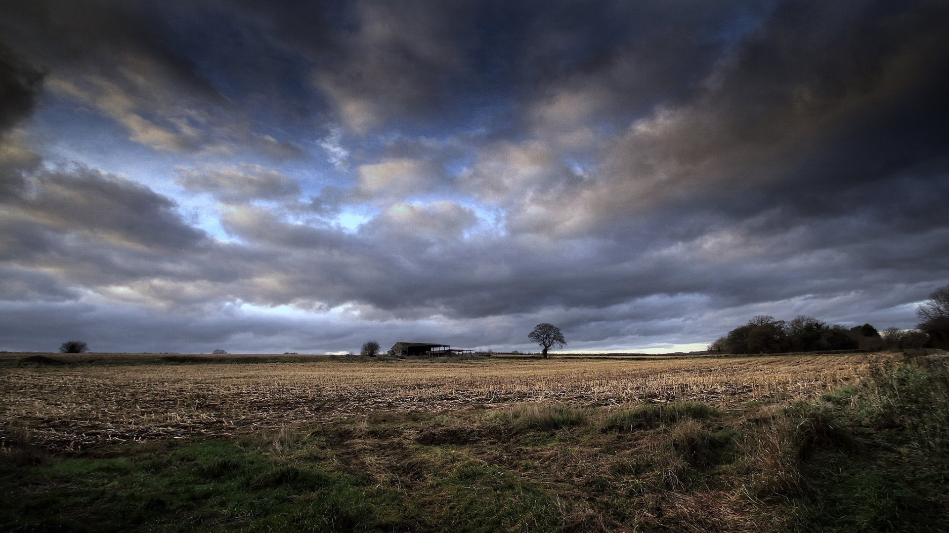 HD PC desktop wallpaper background: distant man-made barn by a lone tree on an open field beneath dramatic storm clouds.