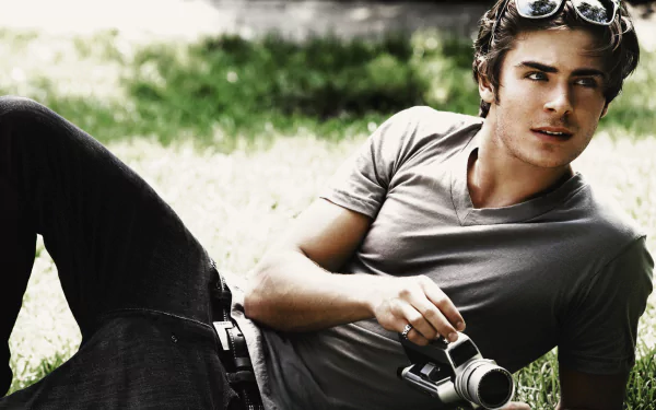 HD desktop wallpaper featuring American actor and celebrity Zac Efron reclining outdoors, holding a vintage camera with a relaxed expression.