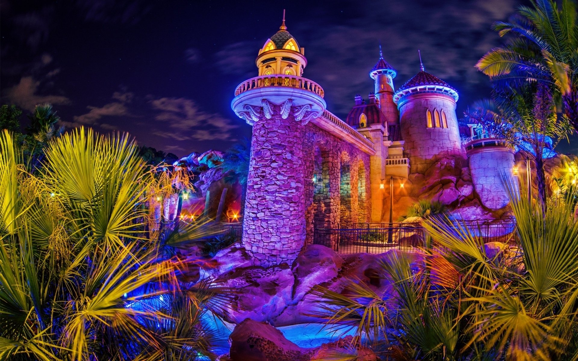 Vibrant nighttime view of a man-made Disney castle at Disneyland, illuminated with colorful lights, framed by lush greenery, captured in HD for a PC desktop wallpaper.