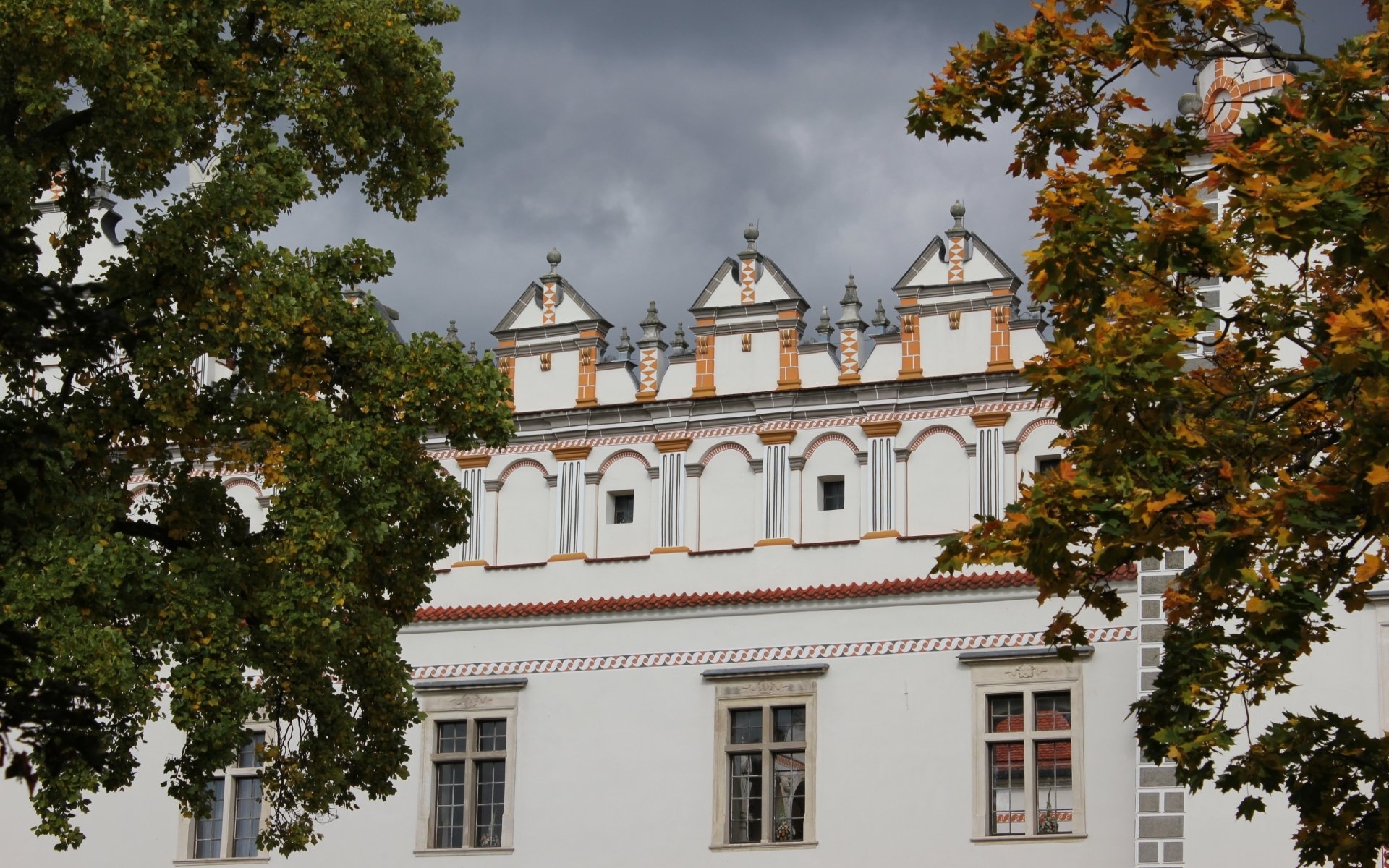 HD desktop wallpaper featuring the man-made Baranów Sandomierski Castle framed by green and autumnal trees under a cloudy sky.