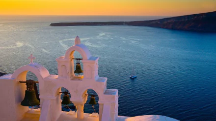 HD desktop wallpaper: sunset panorama of a man-made white church bell tower and bells overlooking the Aegean ocean, beach and Santorini village coast, Greece.