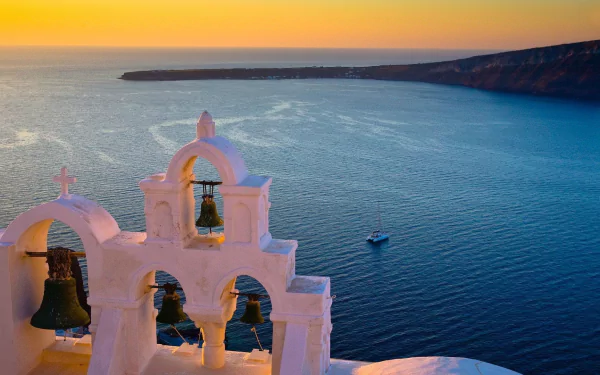 HD desktop wallpaper: sunset panorama of a man-made white church bell tower and bells overlooking the Aegean ocean, beach and Santorini village coast, Greece.