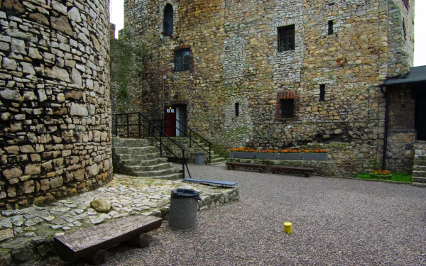 HD PC desktop wallpaper of a man-made Bedzin Castle courtyard: weathered stone walls and arched windows, steps leading up, gravel yard with bench, flowerbed and a small yellow object.