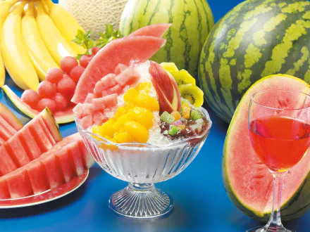 A vibrant dessert bowl filled with colorful fruit, surrounded by watermelons, bananas, and a glass of fruit juice, creating a lively food-themed backdrop for your desktop.