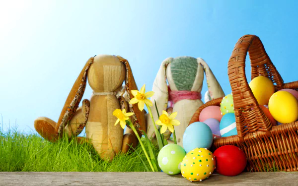 Easter holiday scene with colorful decorated eggs, two bunny plush toys, daffodils, and a wicker basket on grass against a clear blue sky, in 4K Ultra HD resolution.