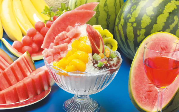 A vibrant dessert bowl filled with colorful fruit, surrounded by watermelons, bananas, and a glass of fruit juice, creating a lively food-themed backdrop for your desktop.