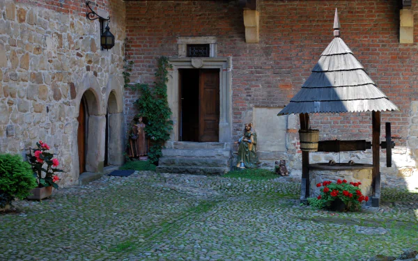 Debno Castle courtyard (man-made): stone walls, arched doorways, wooden well and flower pots on a mossy cobbled ground — HD PC desktop wallpaper/background.