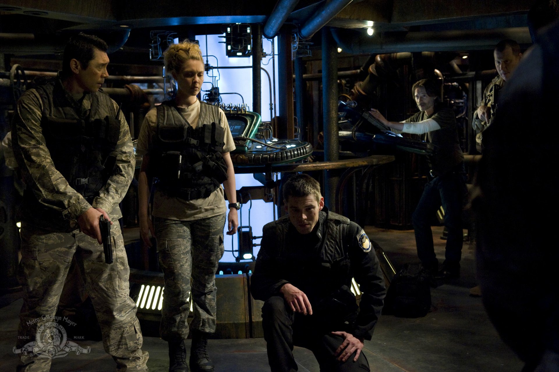 Stargate Universe TV show wallpaper: dimly lit ship interior with uniformed crew gathered around a console, one kneeling, others standing amid pipes and equipment — HD PC desktop background.