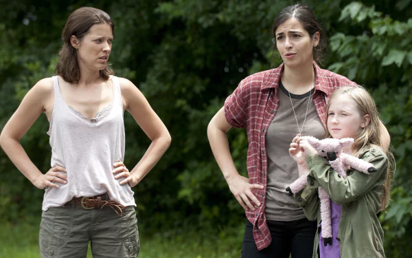 HD PC desktop wallpaper from The Walking Dead showing three women in a wooded setting: two with hands on hips and a young girl clutching a stuffed toy.