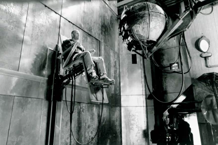 Black-and-white 12 Monkeys (Movie) 2K Quad HD PC desktop wallpaper: a man strapped to a chair in a grim industrial room facing a large suspended mechanical device.