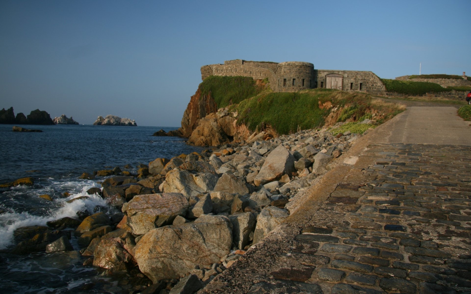HD desktop wallpaper featuring the man-made Fort Clonque perched on rocky cliffs overlooking the calm sea under a clear blue sky.