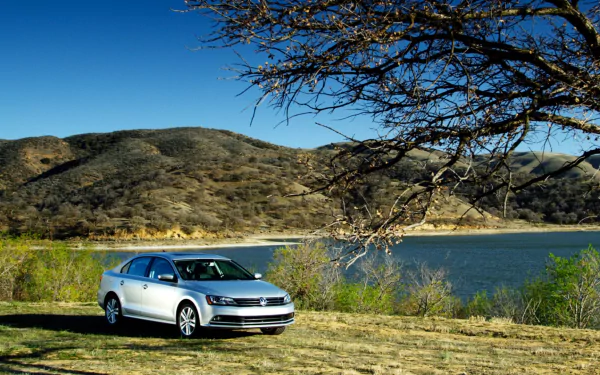 Silver Volkswagen Jetta vehicle by a lakeshore with rolling hills and a tree branch under a clear blue sky — HD PC desktop wallpaper/background.