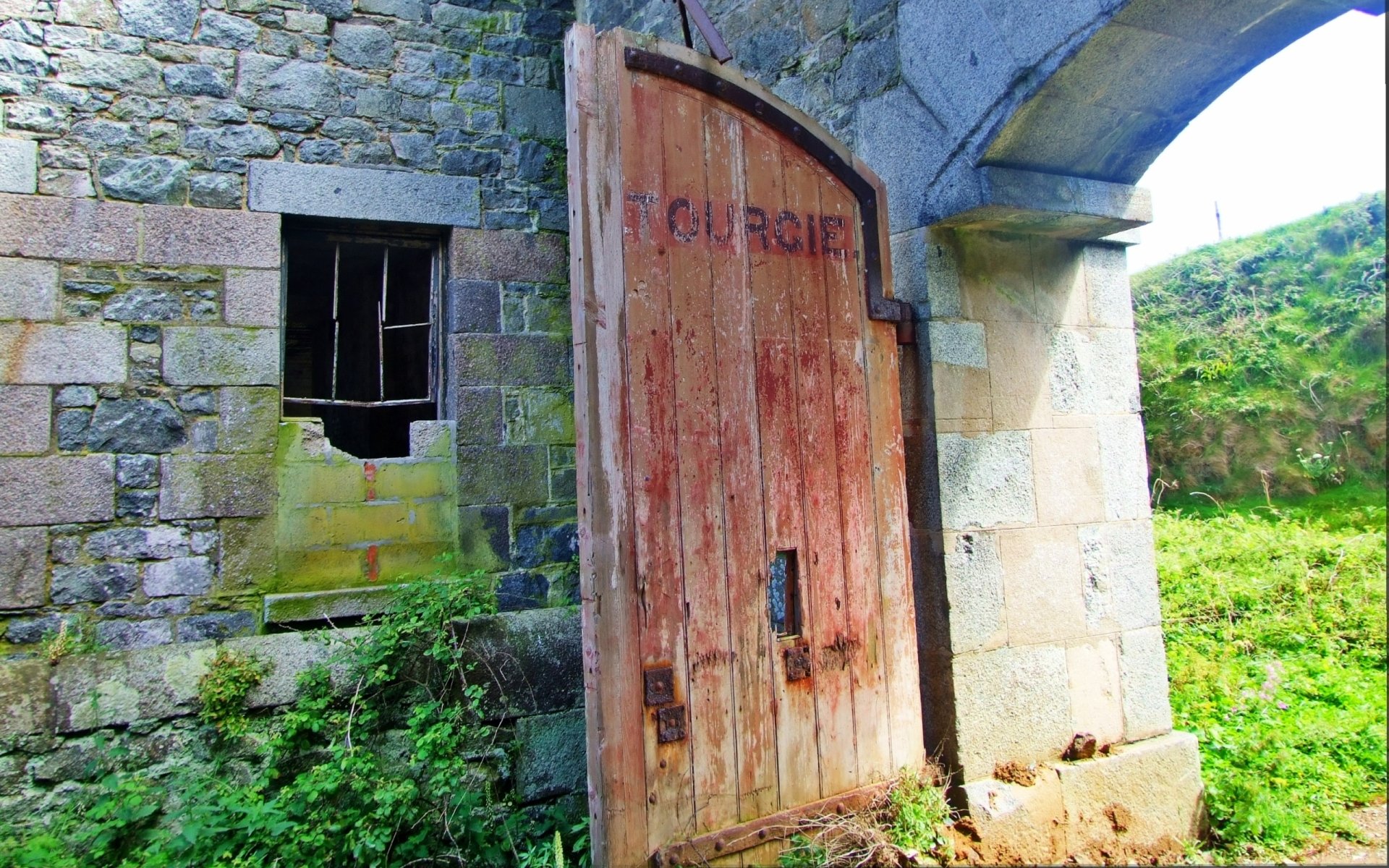 Man-made Fort Tourgis: weathered wooden gate in a stone arch, mossy walls and a grassy slope — 2K Quad HD PC Desktop Wallpaper and Background.