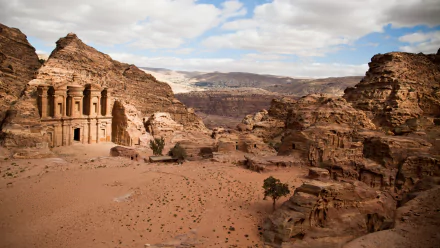 HD desktop wallpaper showcasing the ancient man-made rock architecture of Petra set against a vast desert landscape under a partly cloudy sky.