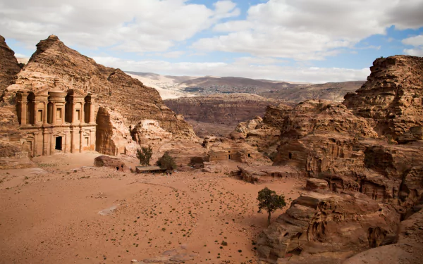 HD desktop wallpaper showcasing the ancient man-made rock architecture of Petra set against a vast desert landscape under a partly cloudy sky.