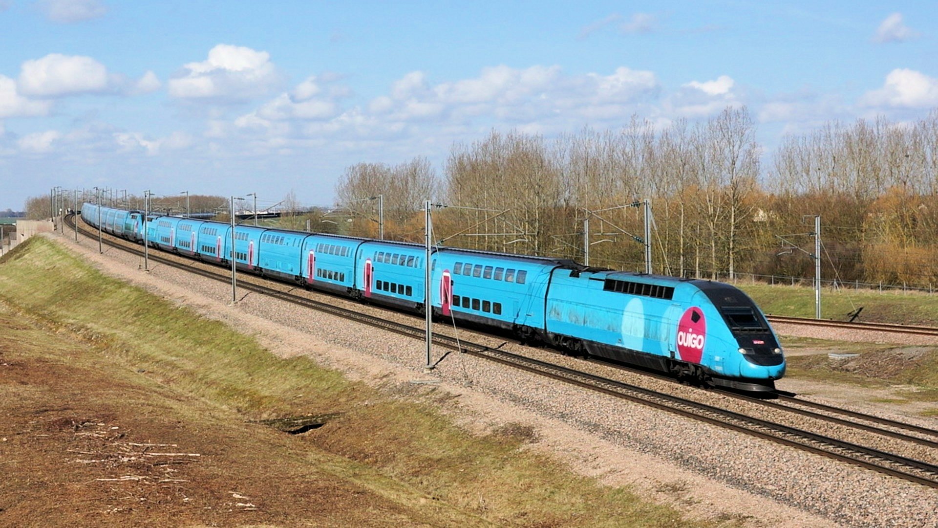 A sleek blue high-speed train glides along tracks under a bright sky, surrounded by earthy tones and distant trees, making for a striking HD desktop wallpaper.