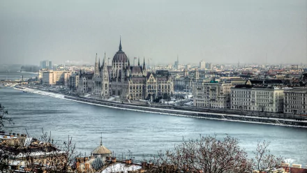 A panoramic view of Budapest, showcasing the iconic Parliament building along the Danube River, surrounded by a winter landscape and urban architecture.