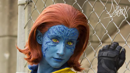 A woman in vibrant blue cosplay, resembling a character with striking yellow eyes and reddish-brown hair, poses confidently against a chain-link fence. HD wallpaper background.