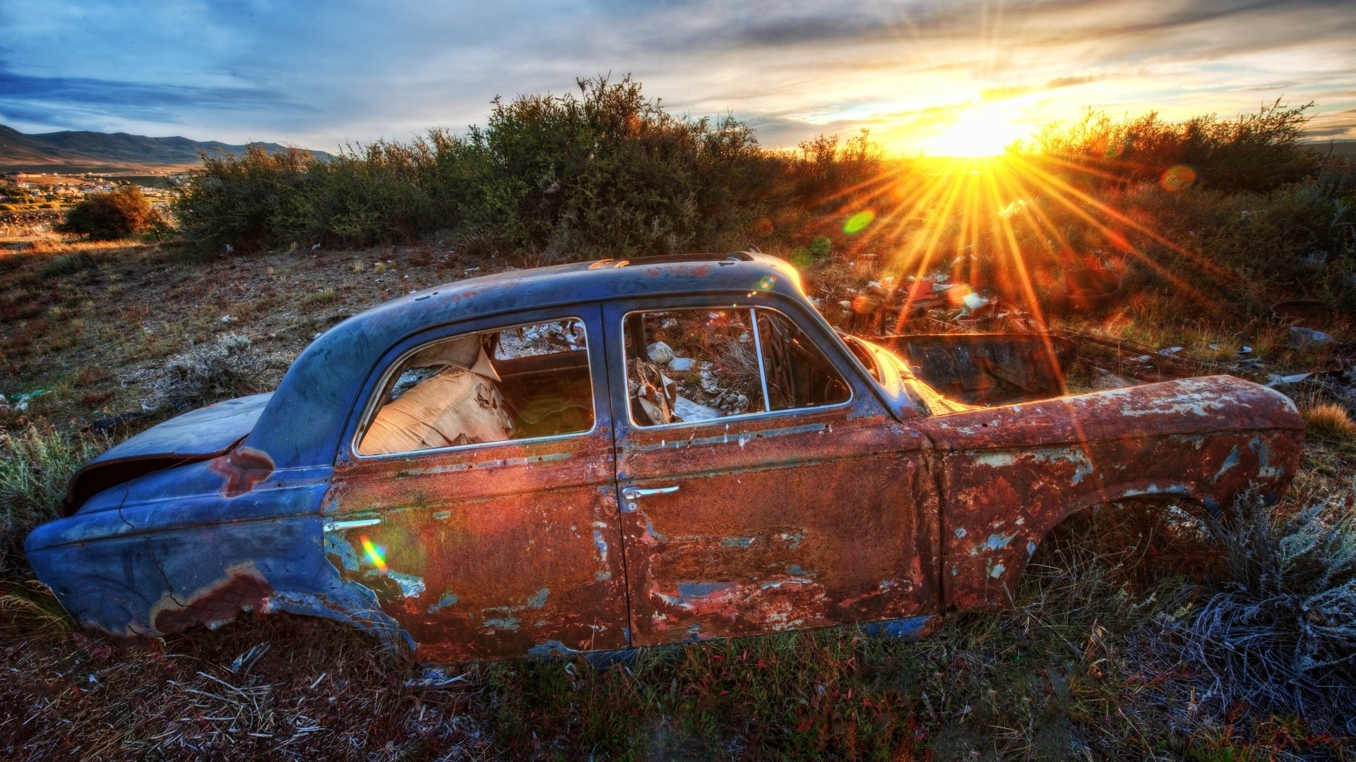 HD desktop wallpaper of a rusted vintage car wreck resting in an overgrown field at sunset, sunburst lighting worn paint and surrounding brush.