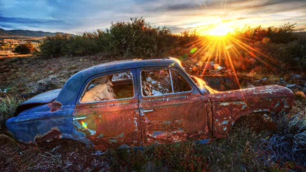 HD desktop wallpaper of a rusted vintage car wreck resting in an overgrown field at sunset, sunburst lighting worn paint and surrounding brush.