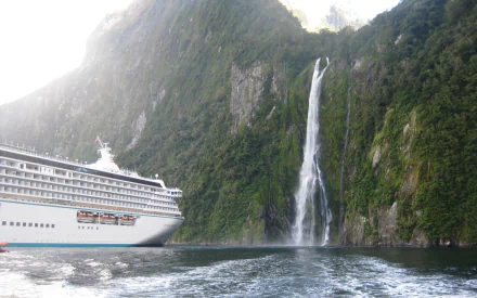 Nature HD PC desktop wallpaper background of Stirling Falls: a tall waterfall cascading beside a cruise ship in a green fjord with calm, reflective water.
