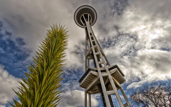 HD desktop wallpaper featuring Seattle's iconic man-made Space Needle against a dramatic cloudy sky with green foliage in the foreground.