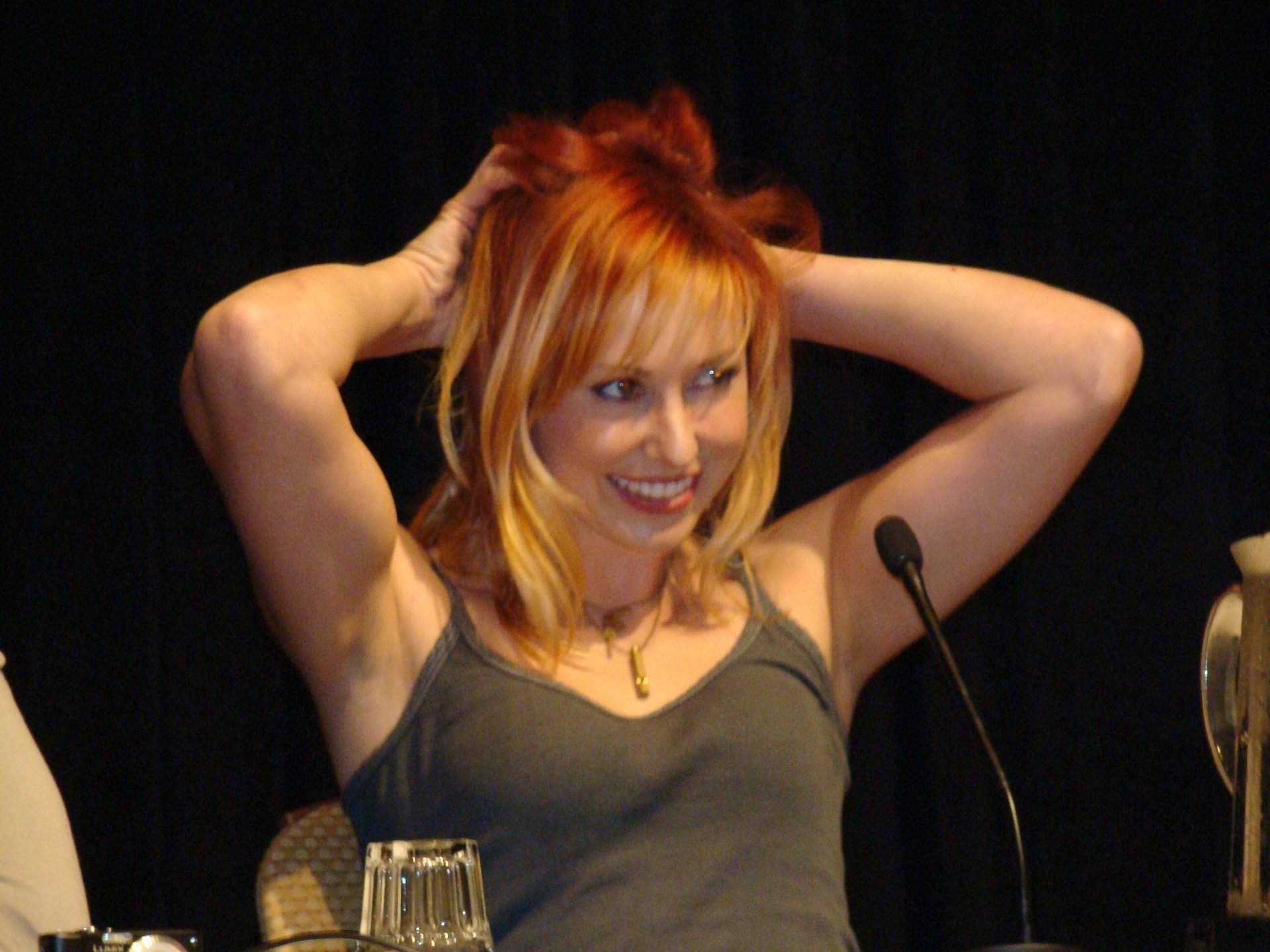 Blonde female celebrity smiling with hands in her hair at a panel microphone — 2K Quad HD PC desktop wallpaper/background.