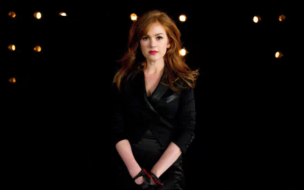 A striking HD desktop wallpaper featuring Australian actress Isla Fisher, with her red hair and stylish black outfit, standing confidently against a dark backdrop illuminated by soft lights.