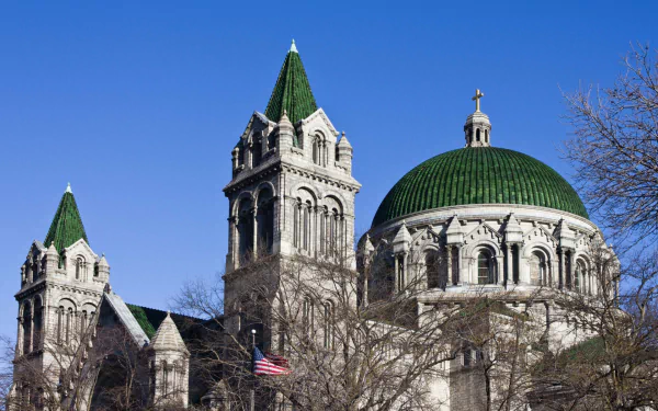 HD PC desktop wallpaper showing the religious Cathedral Basilica of Saint Louis: green domes and spires against a clear blue sky, framed by bare trees and a waving American flag.