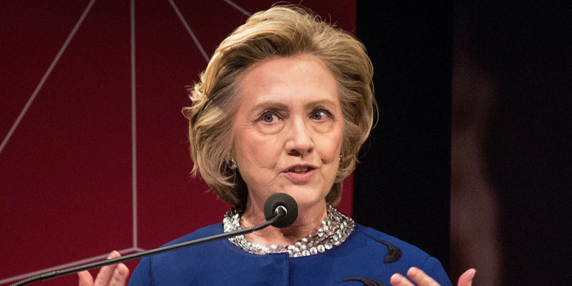 HD desktop wallpaper of a woman speaking at a podium, dressed in blue with a microphone in front.